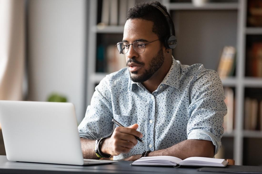 A man at his laptop wearing a headset with a pen in hand.