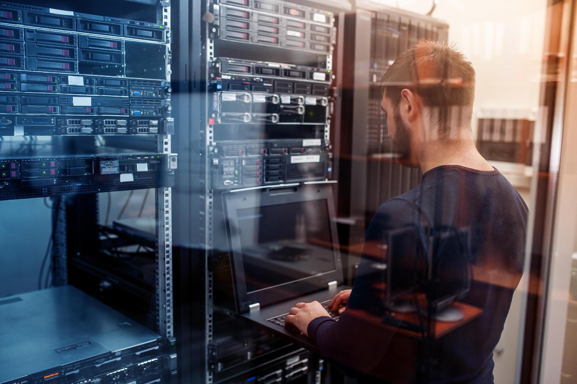 A man types on a black laptop while standing in front of network servers.