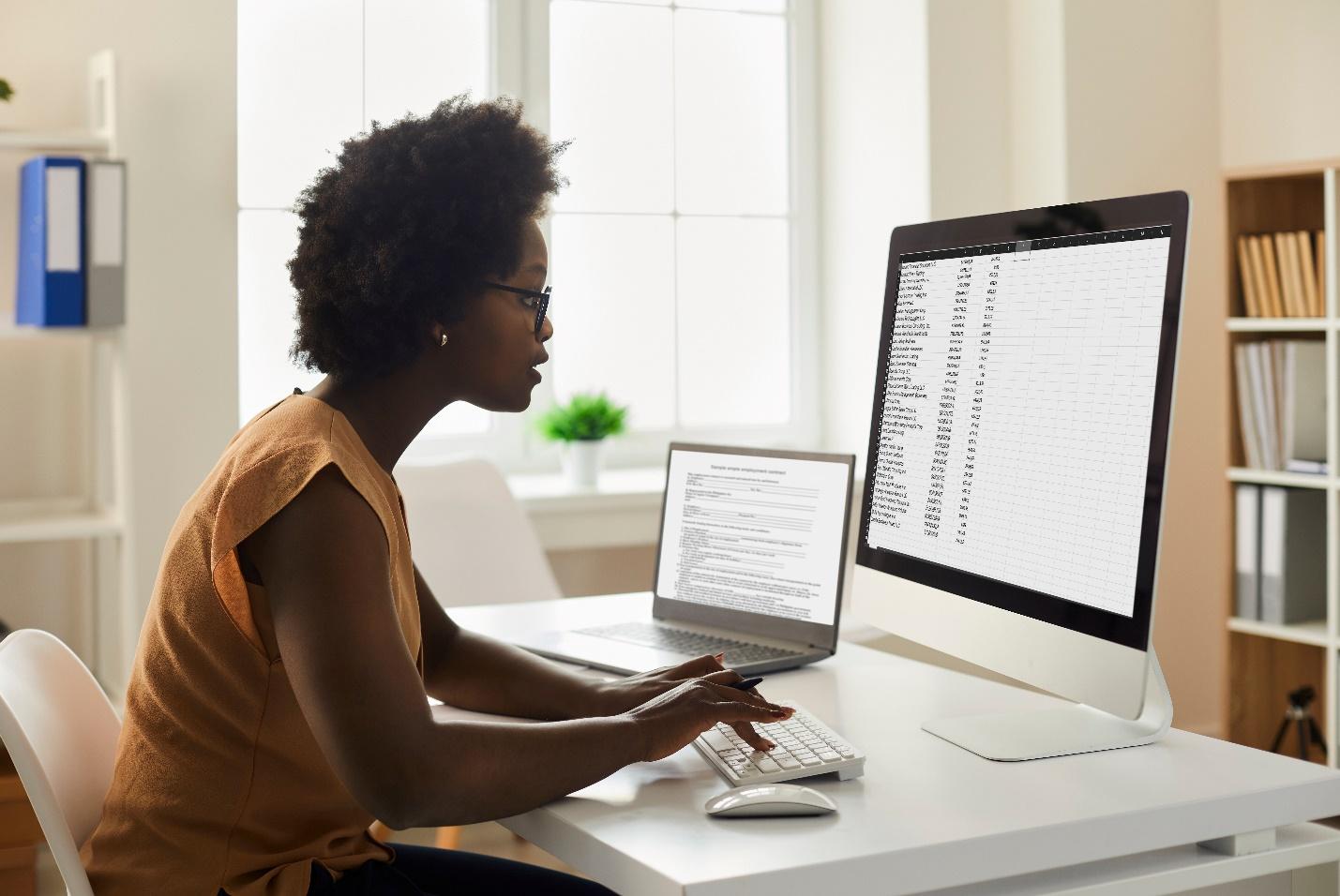 An office employee seated at a desk scrolls through a list on her laptop and a large monitor.