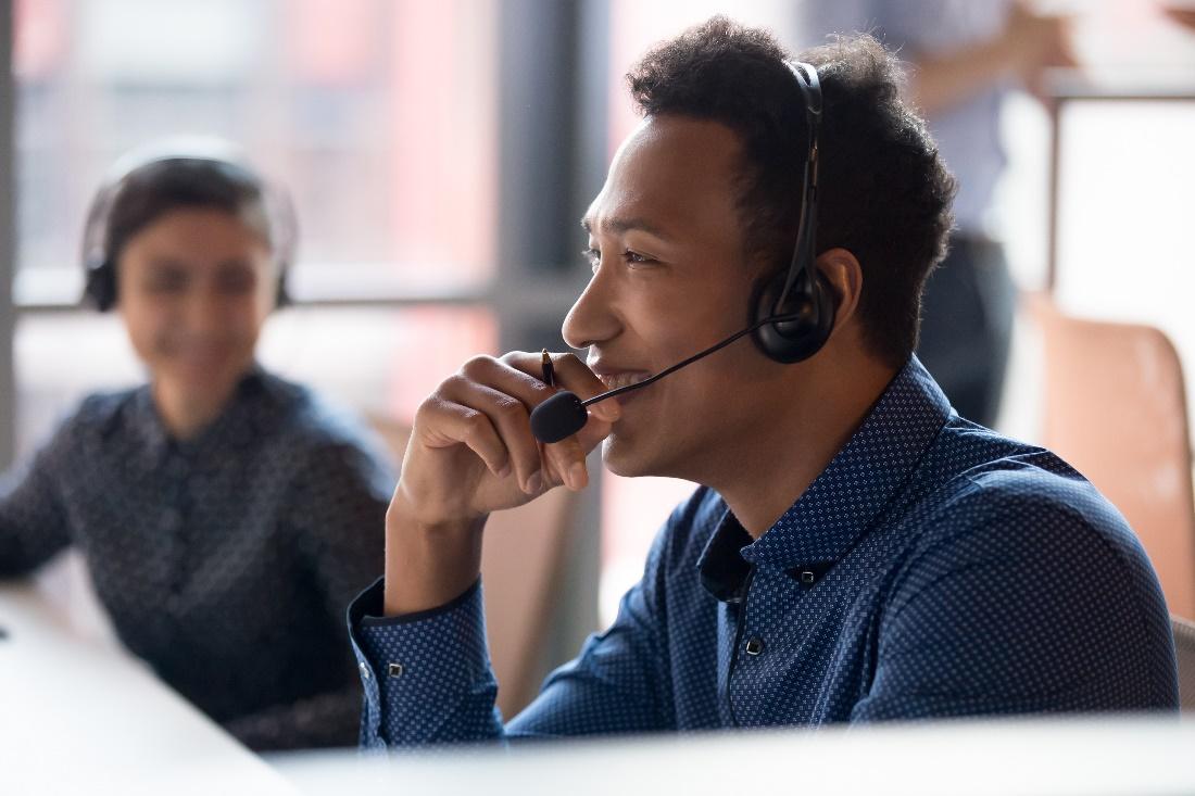 Man talks into a headset with a woman, also wearing a headset, in the background