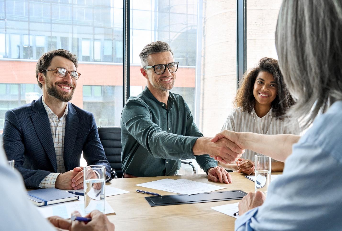 A happy businessperson shakes hands with another professional across a desk while two other professionals look on and smile.