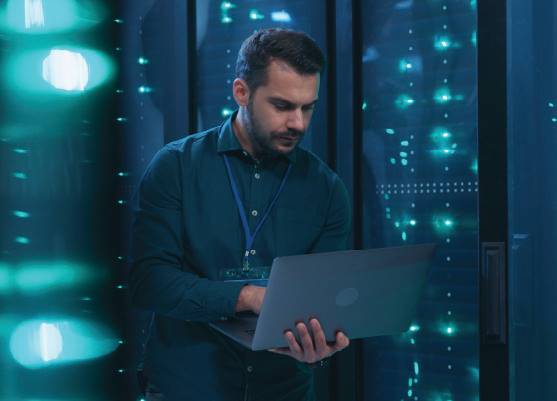 bearded male it technician working on laptop in front of server racks