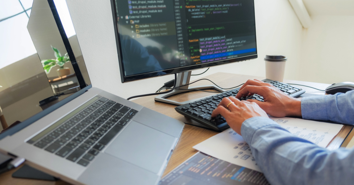 A man sitting at his desktop computer working on code, with a laptop computer to his left.