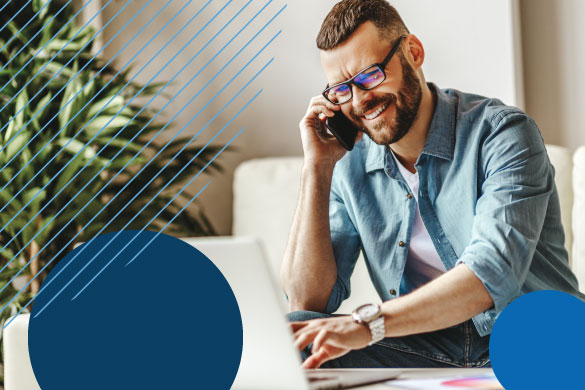 White man wearing glasses and a denim shirt is walking on the phone while using a computer. He is smiling